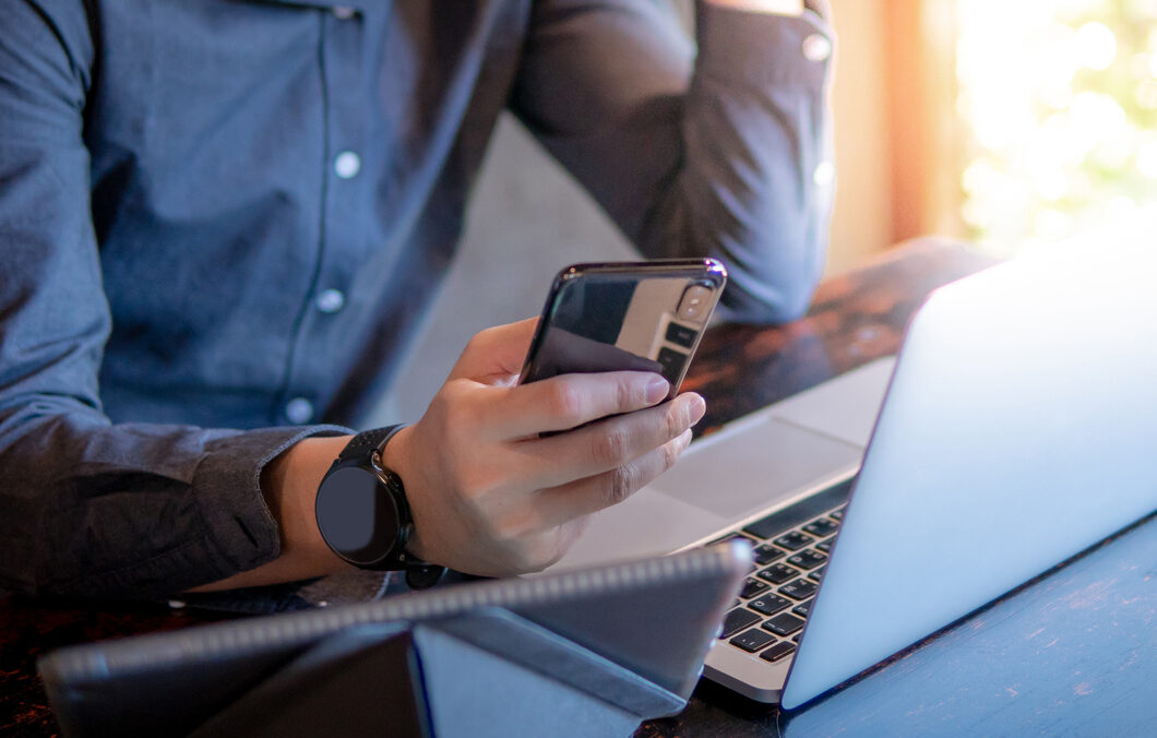 Male hand holding smartphone. Businessman using laptop computer and digital tablet while working in the cafe. Mobile app or internet of things concepts. Modern lifestyle in digital age.
